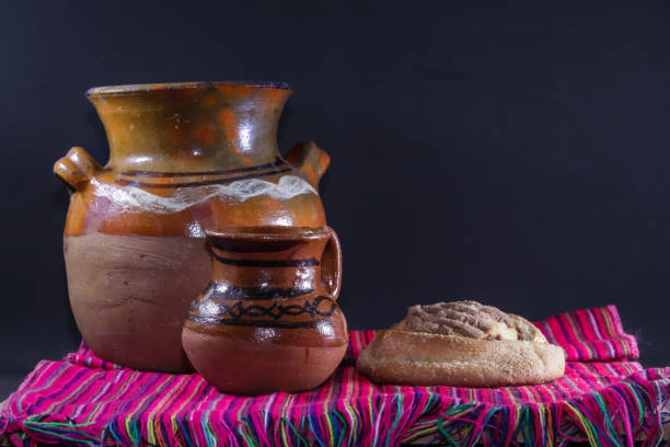 Coffee pot, next to clay cup accompanied with a sweet bread on a Mexican rebozo of various colors with a black background