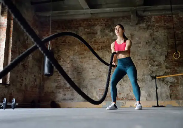 Photo of Young woman exercising with battle ropes at the gym. Strong female athlete doing gym workout with battle rope