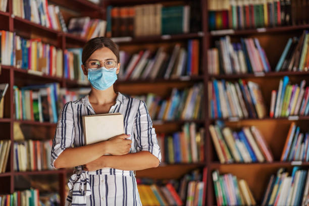 portret młodej atrakcyjnej dziewczyny z college'u stojącej w bibliotece z maską na trzymaniu książki. badanie podczas koncepcji wirusa korony. - book reading dress women zdjęcia i obrazy z banku zdjęć