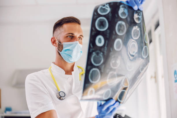 attraente giovane medico dedicato in piedi in ospedale con maschera facciale e guanti di gomma e guardando le radiografie del cervello del paziente. - doctor brain x ray x ray image foto e immagini stock