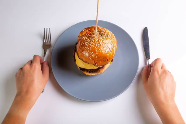 Homemade cheeseburger on a gray plate and a person with cutlery in his hands on a white background. Hands in the frame. View from above. Horizontal orientation. High quality photo