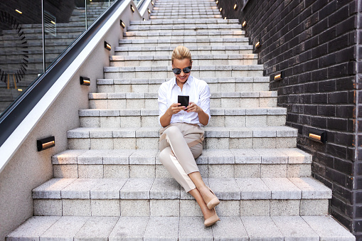 Attractive glamorous smiling positive blond fashionable woman sitting on the stairs outdoors and using smart phone.