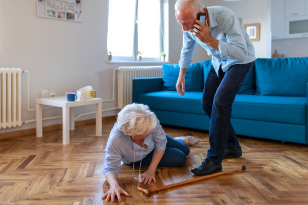 Disabled Elderly Woman had a Misfortune and Fell on the Wooden Floor in the Living Room of her Apartment. Senior wife sitting on floor while retired husband talking on smartphone calling an ambulance grey hair on floor stock pictures, royalty-free photos & images