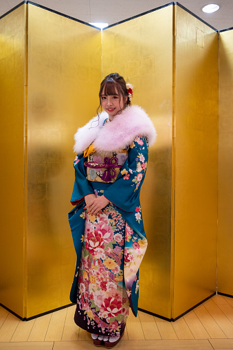 Full length portrait of young woman in ‘Furisode’ kimono with fur neck stole standing in front of golden ‘Byobu’ folding screen for ‘Seijin Shiki’ coming-of-age ceremony