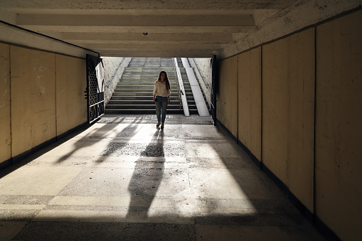 Young woman descends into gloomy underpass. Two shadows from people descend behind her. Concept of follow, defenseless and danger in desolate place