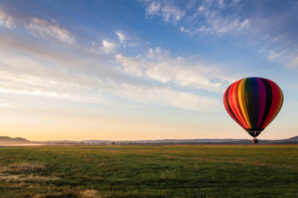 Hot Air Balloon in colorful rainbow stripes begins ascent over farm field as sun rises blue cloudy sky stock photo