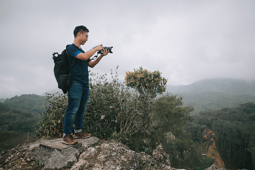 asian chinese male hiker exploring mossy forest in cameron highland sitting on rock taking photo with his DSLR