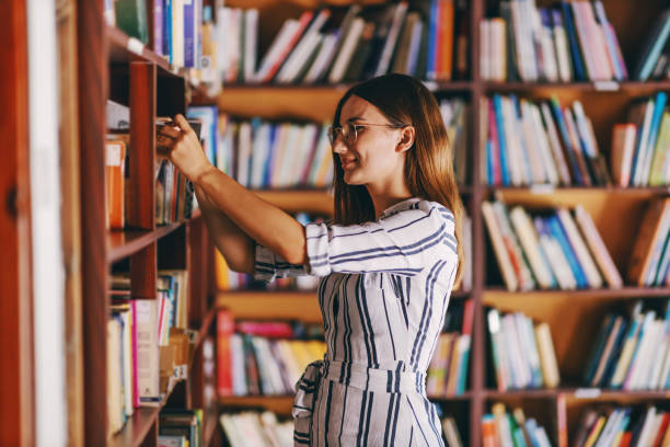 젊은 화려한 여학생은 선반을 예약옆에 서서 시험을 위해 책을 찾고 있습니다. - librarian 뉴스 사진 이미지