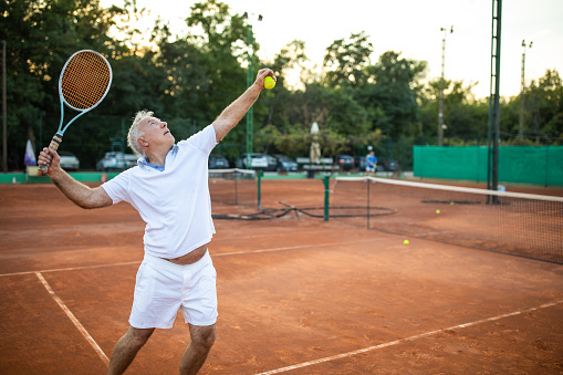 Senior man serving on tennis court during summer day