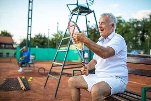 Senior tennis player resting on bench beside tennis court during match