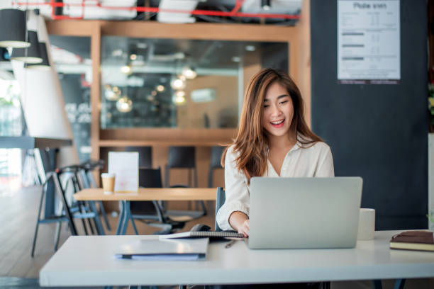 happy young asian businesswoman sitting on her workplace in the office. young woman working at laptop in the office. - asia imagens e fotografias de stock