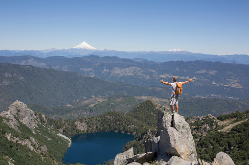 Hiker stands on rock pinnacle above lake and snowcapped volcano