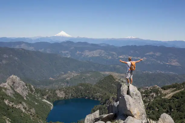 Photo of Hiker stands on rock pinnacle above lake and snowcapped volcano