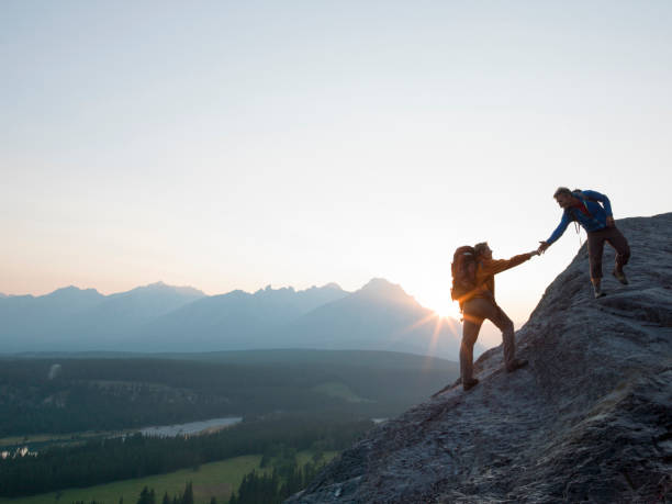 zwei bergsteiger bieten helfende hand auf einem felsrücken bei sonnenaufgang über einem tal - berggipfel stock-fotos und bilder