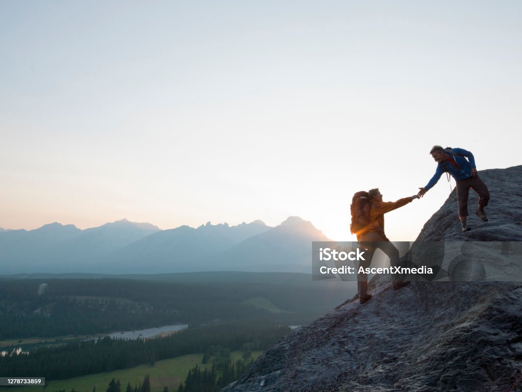 Two mountaineers offer helping hand on a rock ridge at sunrise above a valley The sun is rising over the distant peaks Mountain Climbing Stock Photo