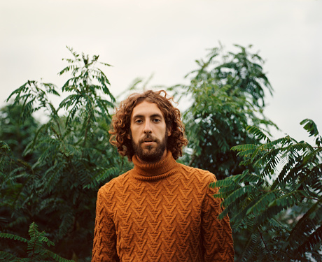 Portrait of  young Caucasian man with curly hair in orange sweater on the background of green leaves