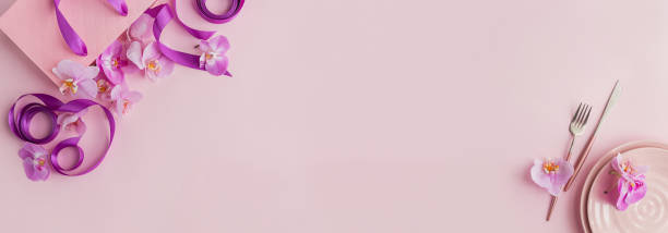 istock Flower and table settings overhead composition on light pink background. Pink ceramic plates, cutlery, pink gift bag with purple ribbons and pink orchid flowers. Holiday modern table decoration 1278742865