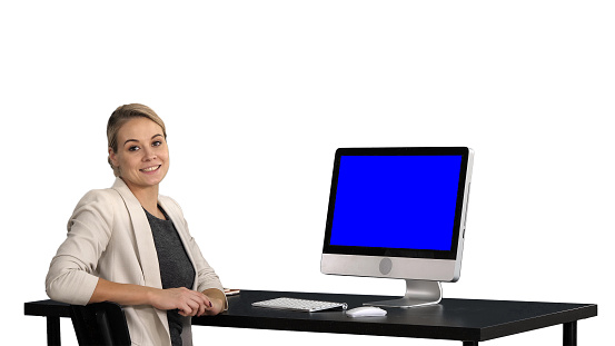 View to the screen of computer. Confident focused businesswoman talking to camera and looking to monitor near her, white background. Blue Screen Mock-up Display. Professional shot in 4K resolution. 007. You can use it e.g. in your commercial video, business, presentation, broadcast
