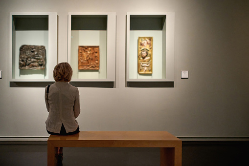 Young Caucasian woman reading through a digital tablet with data of the displayed objects while her biracial husband is checking out the stone remains of an Ancient Roman town. Candid shot over the display cases in the middle of museum gallery.