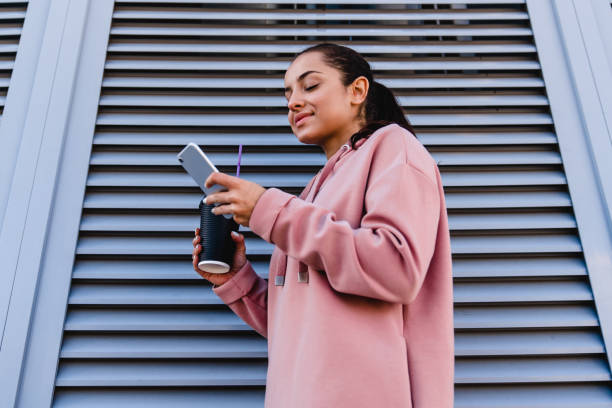 jovem garota esportiva está usando seu telefone inteligente segurando uma xícara de café perto da parede cinza - fresh coffee audio - fotografias e filmes do acervo