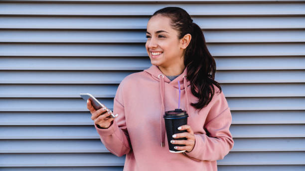 feche a foto de uma jovem linda segurando seu telefone e café em frente à parede de tapumes - fresh coffee audio - fotografias e filmes do acervo
