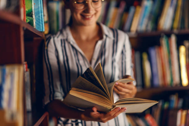 joven joven sonriendo atractiva universitaria apoyada en los estantes de los libros en la biblioteca y leyendo un libro. - professor librarian university library fotografías e imágenes de stock