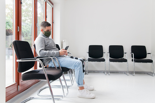 Joven con máscara facial sentado en una sala de espera de un hospital u oficina mirando el teléfono inteligente photo