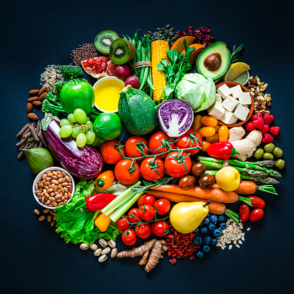 Vegan food: large group of multicolored fresh organic fruits, vegetables, nuts and spices arranged in a circle shot from above on dark background. The composition includes green apple, kiwi, pomegranate, lemon, orange slices, peach, fig, pear, grape, berries, almonds, pistachio, oat flakes, pumpkin seeds, flax seeds, rice, dried herbal tea, olives, olive oil, goji berries, pinto beans, wholegrain pasta, celery, tomatoes, carrot, lettuce, eggplant, avocado, broccoli, ginger, pepper, turmeric, asparagus, green beans, corn, tofu, among others. High resolution 28Mp studio digital capture taken with SONY A7rII and Zeiss Batis 40mm F2.0 CF lens