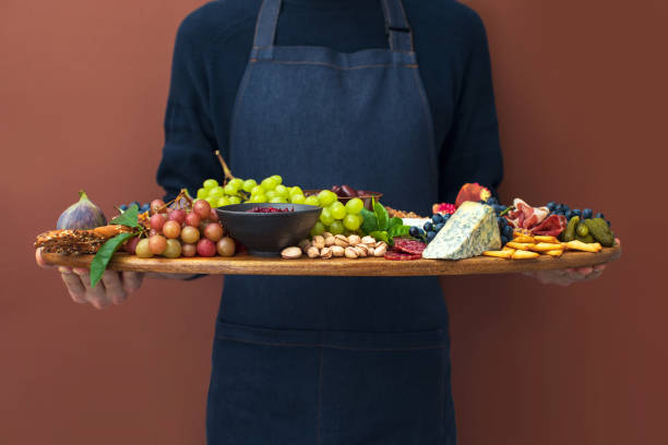 Man in apron holding charcuterie board, front view Man in an apron serving charcuterie board, front view of meat and cheese plateau with fruits snacks, and olives fusion food stock pictures, royalty-free photos & images