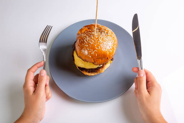 homemade cheeseburger on a gray plate and a man with cutlery in his hands on a white background. hands in the frame. view from above. horizontal orientation. high quality photo - buns of steel imagens e fotografias de stock