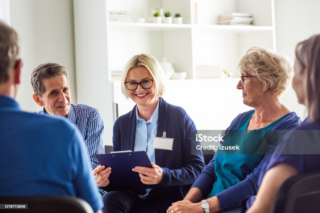 Cheerful psychotherapist talking with senior people Senior people sharing issues with patients and coach during meeting in nursing home. Mental health professional is sitting with people at community center. They are discussing about mental wellbeing. Meeting Stock Photo