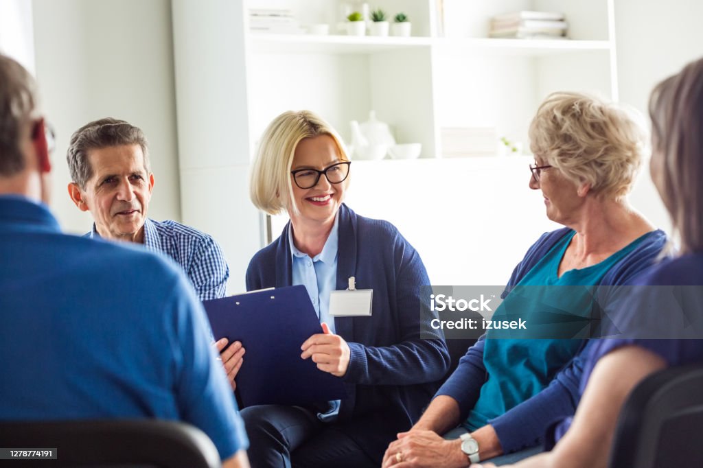 Cheerful psychotherapist talking with senior people Senior people sharing issues with patients and coach during meeting in nursing home. Mental health professional is sitting with people at community center. They are discussing about mental wellbeing. 70-79 Years Stock Photo