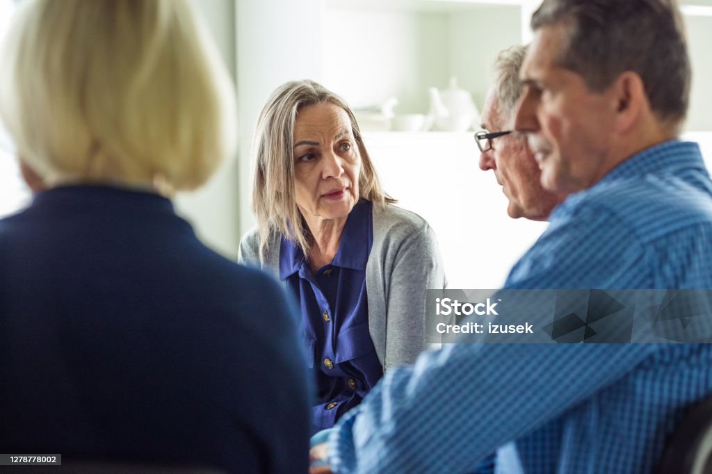 Worried eldery lady during group therapy Senior people sharing issues with patients and coach during meeting in nursing home. Mental health professional is sitting with people at community center. They are discussing about mental wellbeing. Discussion Stock Photo