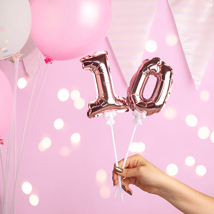 Decoration for birthday party. Female hand holding golden balloon in form of 10 number on pink background. Sparkles wallpaper decor.