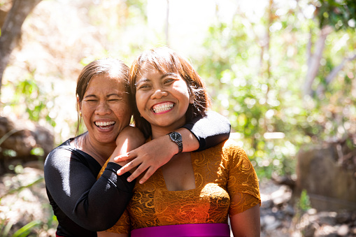 A shot of a two mid adult Balinese sisters wearing traditional clothing, smiling and laughing away from camera. They are standing in a rural area surrounded by forest.
