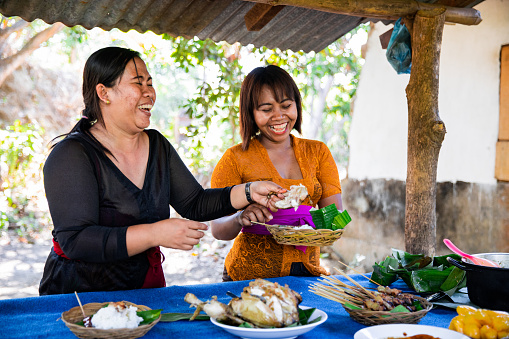 A shot of two Balinese women serving a buffet on a large table under a shelter. They are wearing traditional clothing and are laughing while serving up the food.