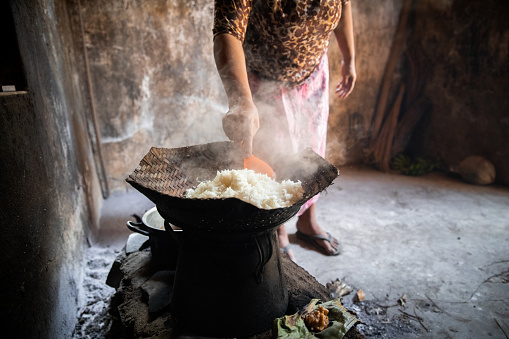 A shot of an unrecognisable Balinese woman standing in in a very basic hut. She is wearing traditional clothing and is steaming rice over an open fire.