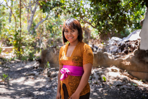 A shot of a mid adult Balinese woman wearing traditional clothing and smiling at the camera. She is standing in front of a hut in a rural area, surrounded by forest.