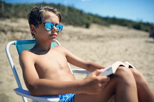 Little boy sitting on a beach, reading a book and sunbathing. The boy is wearing sunglasses. 
Sunny summer day.
Nikon D850