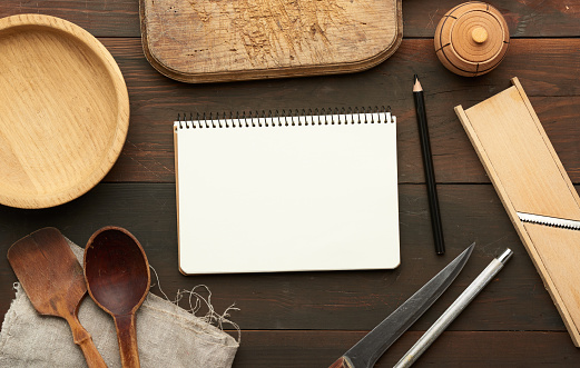 open notebook with blank white sheets and kitchen utensils on brown wooden table, top view