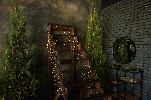 een bruine houten trap met slingers van heldere lichten bevindt zich tegen een grungy grijze concrete muur. kerstboom in de loft. er is een ronde spiegel aan de bakstenen kant met een zwarte plank eronder. - christmas tree stockfoto's en -beelden