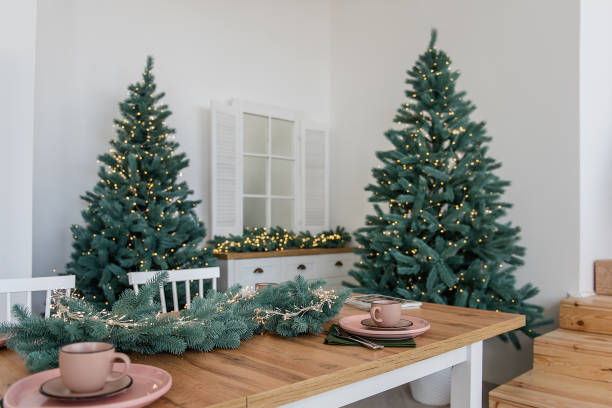 elegante cucina bianca in arredamento, stile alla moda scandinavo. un tavolo in legno ecologico, piatti rosa ceramica, un ramo di albero di natale con una gilanda di luci. copiare lo spazio. modello di capodanno, web card - christmas tree christmas tree artificial foto e immagini stock