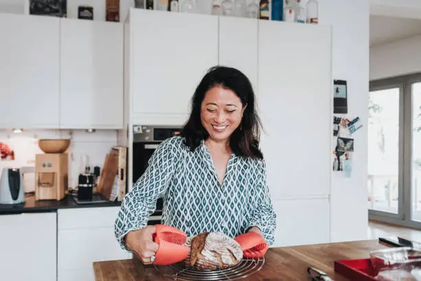 smiling mid aged woman with red oven clothes putting home made warm bread on kitchen table