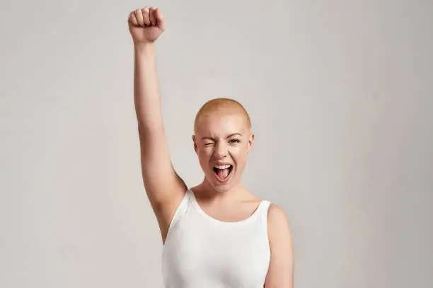 Portrait of a beautiful young caucasian woman with shaved head wearing white shirt, winking at camera and raising clenched fist, standing isolated over grey background. Front view. Horizontal shot