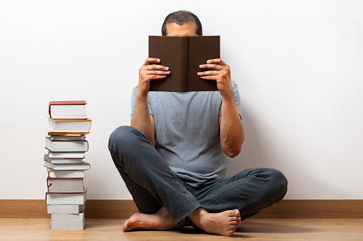 Man sitting on a floor and holding book in front of face at home