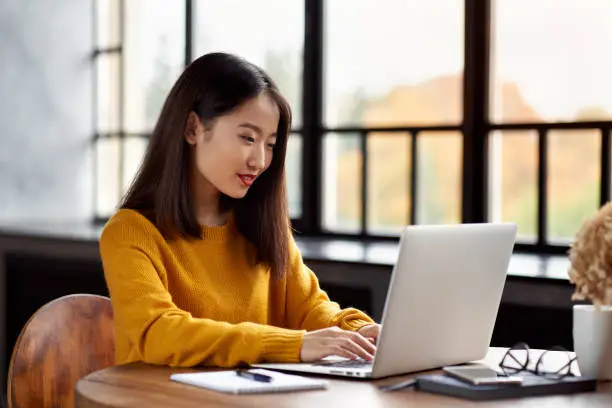 Asian woman working on laptop at home or in cafe. Young lady in bright yellow jumper is sitting at desk typing on computer. Business Asian female in front of window