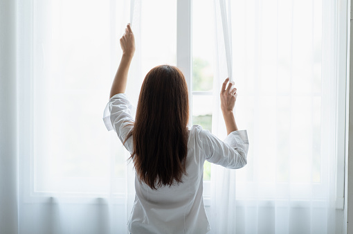 Portrait of young Asian woman opening the curtain in the morning after wake up