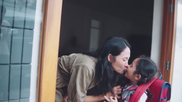 Mother kissing her daughter on the first day of preschool at the door house.