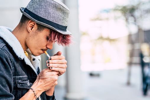 Young modern caucasian teenager male with violet hair and hat light a cigarette- smoking and smoke people concept - teen portrait in real life activity in the city - defocused background