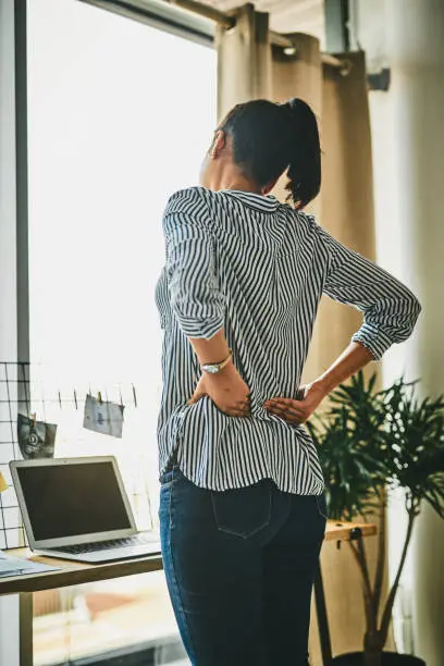 Rearview shot of a young woman suffering with back pain while working from home